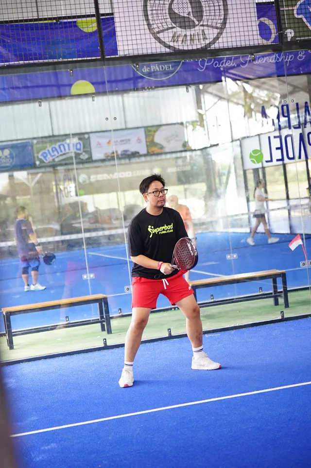 A padel player holding a racket waits on the blue court.